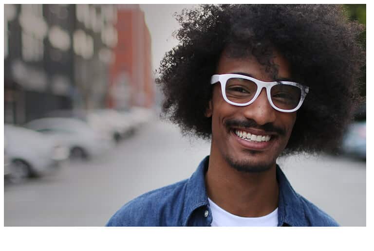 Smiling man with black afro and goatee stands on a street wearing white square glasses #124130 with a chambray shirt and white tee.