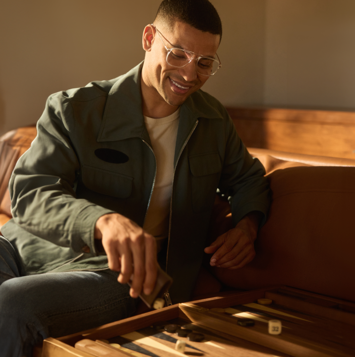 A man wearing Zenni Progressive glasses smiles while playing backgammon, sitting in a warmly lit, cozy room.