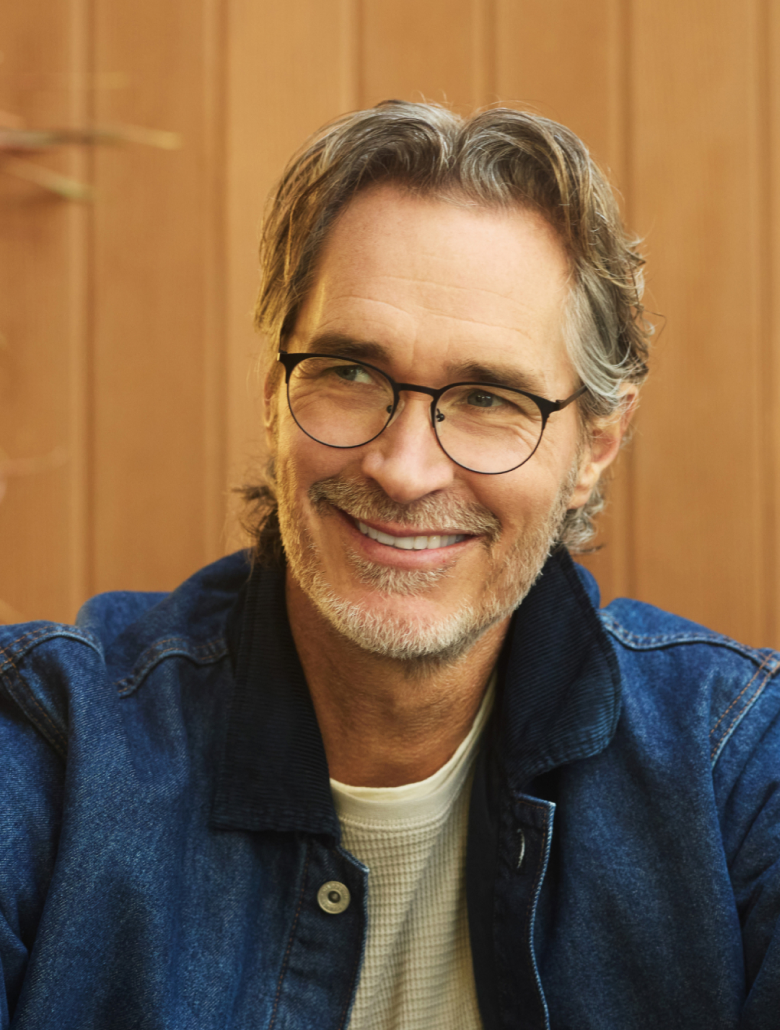 Cheerful middle-aged man with grey stubble, wearing Zenni Progressive glasses and a denim jacket, smiling off-camera in front of a cozy wooden background.