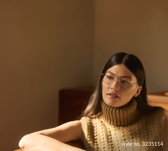 Thoughtful young woman with Zenni Progressive glasses looking away pensively, wearing a chunky turtleneck sweater, in a warmly-lit room.