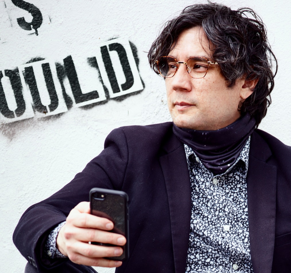 Image of a man wearing a suit, turtleneck shirt, and Zenni glasses. He is holding a cell phone and standing in front of a graffiti wall.
