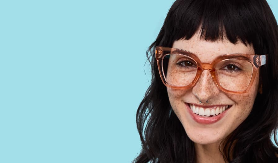 Mujer de cabello largo y oscuro y pecas, sonriendo radiante con unas lentes translúcidas marrones sobredimensionadas, sobre un fondo azul pálido.