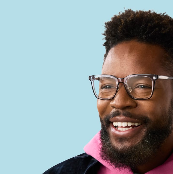 Man with a full beard and bright smile wears clear gray glasses and a pink hoodie, exuding warmth and style.