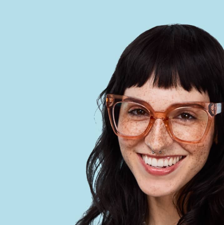 Woman with long dark hair and freckles, smiling brightly in oversized brown translucent glasses, against a pale blue background.
