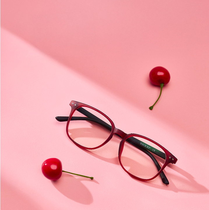 A pair of red eyeglasses with black arms lies on a pink surface surrounded by three cherries.