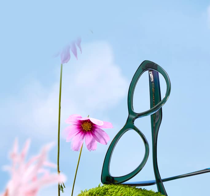 Dark green cat-eye Zenni glasses on a mossy hill and blue sky background with pink flowers.
