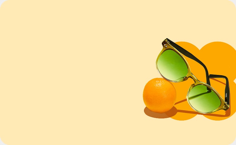 Green round sunglasses with green lenses on an orange background, resting on a small orange.