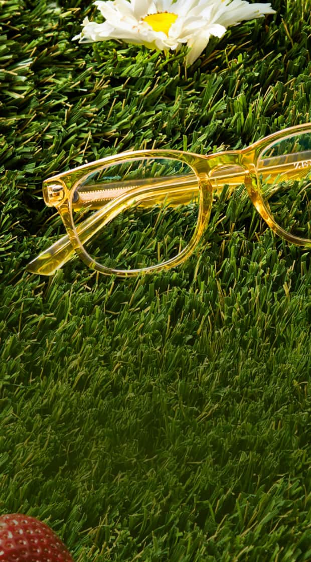 Transparent yellow eyeglasses resting on vibrant green grass, accompanied by a white daisy and a single ripe strawberry.