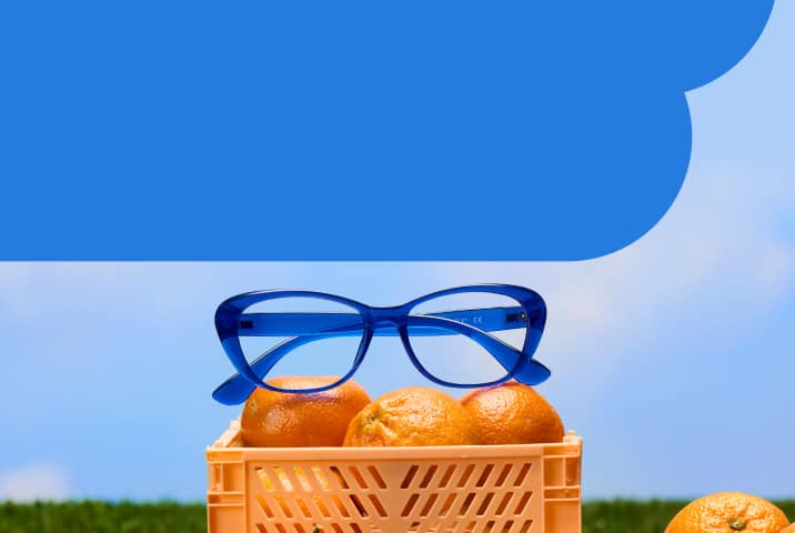A pair of vibrant blue cat-eye glasses resting on top of a crate of oranges in a field of green grass under a clear blue sky.