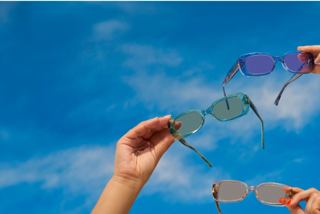 Hands holding pairs of purple, green, and beige rectangle sunglasses up to the blue sky.