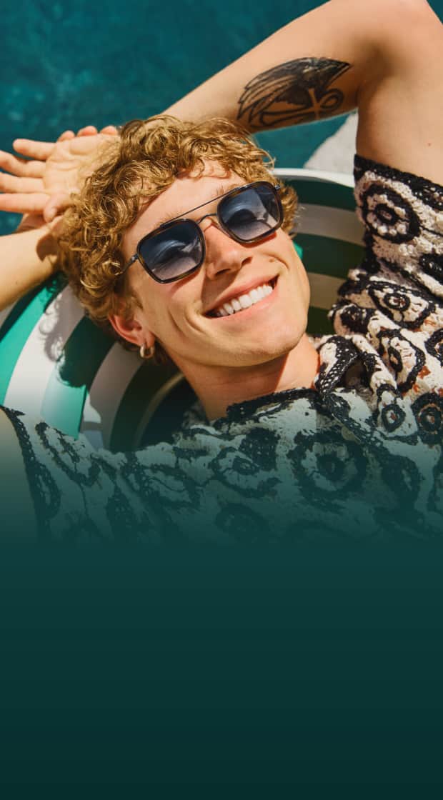 A man with blond curly hair wearing Zenni metal aviator sunglasses with gradient lenses relaxes on an inner tube by a pool.