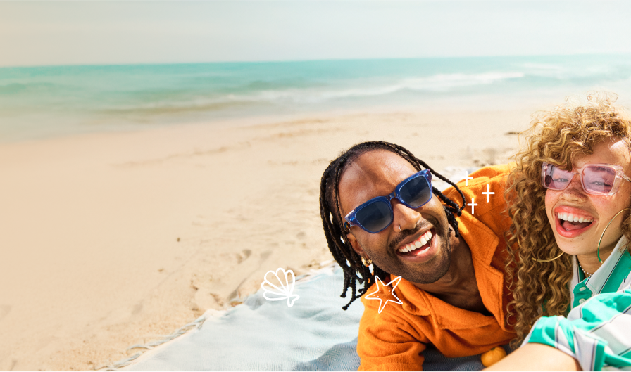 Two people smiling and posing together on a sunny beach, both wearing colorful sunglasses and bright clothing, with the ocean in the background.