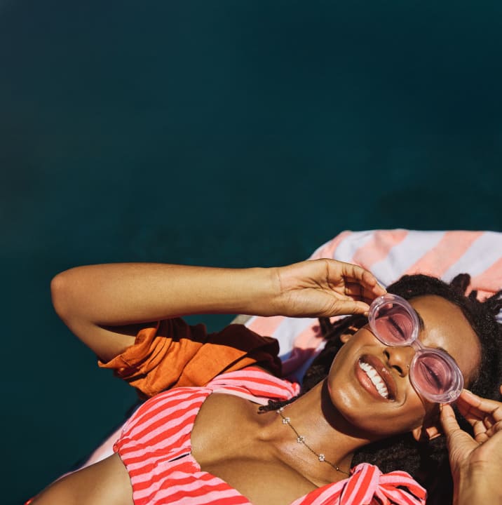 A woman lies on a striped towel by a dark teal pool, smiling and adjusting their large pink sunglasses. She wears a bright pink and orange outfit with a delicate necklace, and sunlight highlights the colorful look.