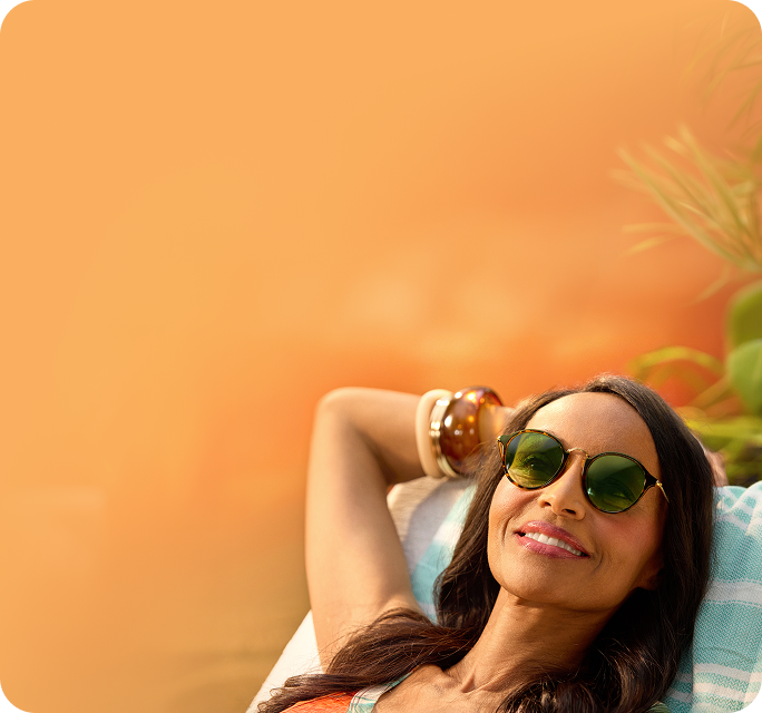 A smiling woman with long dark hair relaxes on a turquoise and white striped towel, wearing round tortoiseshell sunglasses with green-tinted lenses against a warm, golden sunset background.