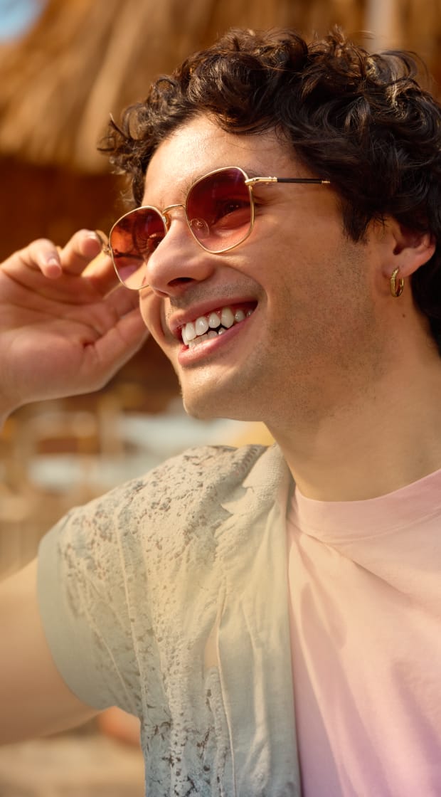 Five diverse people in colorful clothing smile and laugh while showcasing a variety of eyewear including gold aviators, rose gold cat-eye frames, and polka-dot glasses against a light blue background.