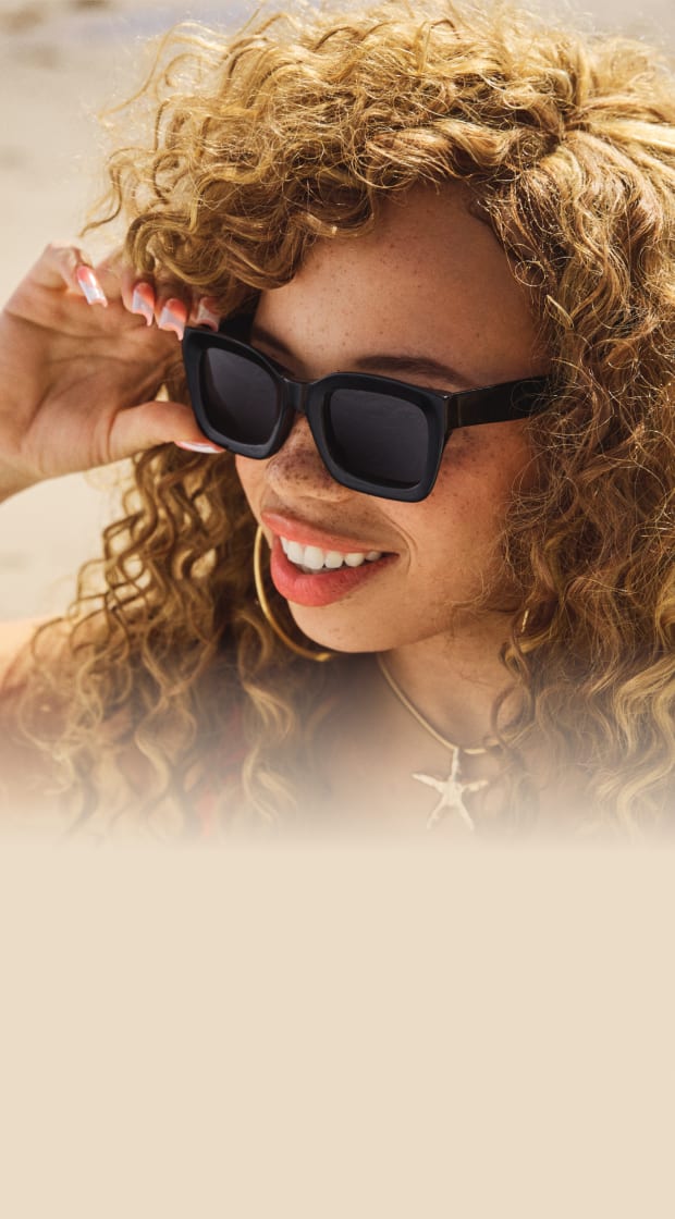 Smiling woman with curly blonde hair wearing black square sunglasses, adjusted with her hand, set against a sandy beach background.
