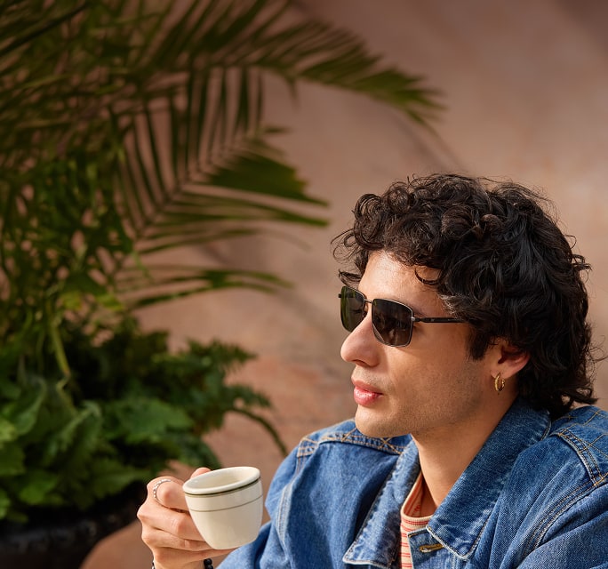 A man in black rectangle Zenni sunglasses. He has mid-length curly brown hair and is sipping an espresso.