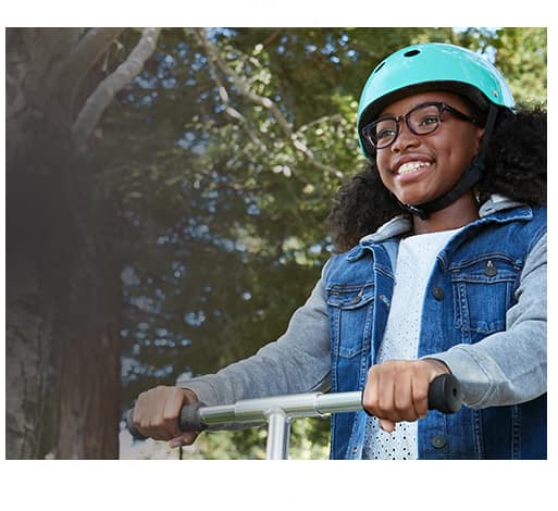 Young girl smiles while riding a scooter outside wearing medium-sized Dare square glasses #4440616 in blue tortoiseshell from the Style Squad kids’ collection with Blokz Trivex lenses and light blue helmet.