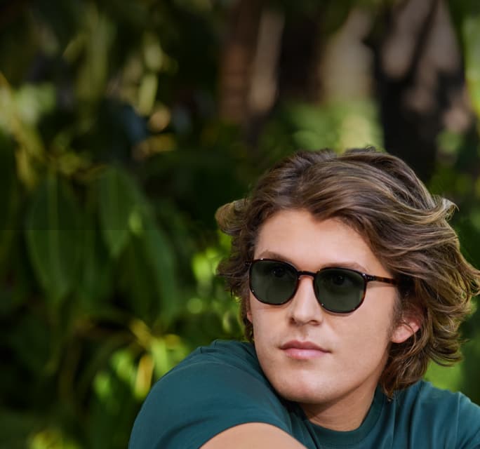Close-up of a man with wavy brown hair looking to the side, wearing tortoiseshell glasses with dark lenses, set against a blurry background of green foliage.