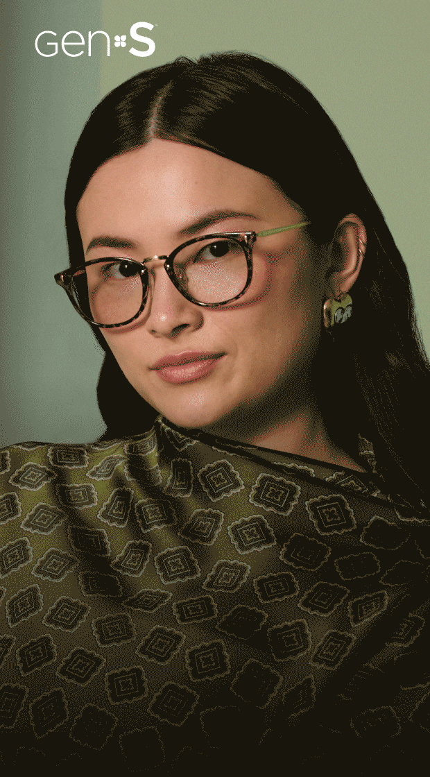 Animated GIF of a woman wearing tortoiseshell eyeglasses with bright green temples and a patterned green scarf, demonstrating photochromic Gen S lenses shifting back and forth between dark tinted sunglasses and clear prescription lenses against a sage green background.