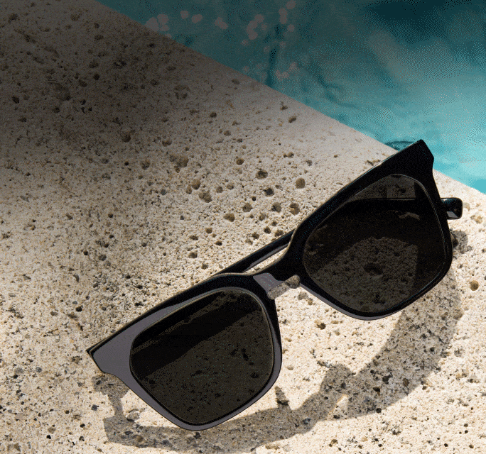 Black square sunglasses with dark tint lenses resting on a textured poolside stone ledge above shimmering blue water.