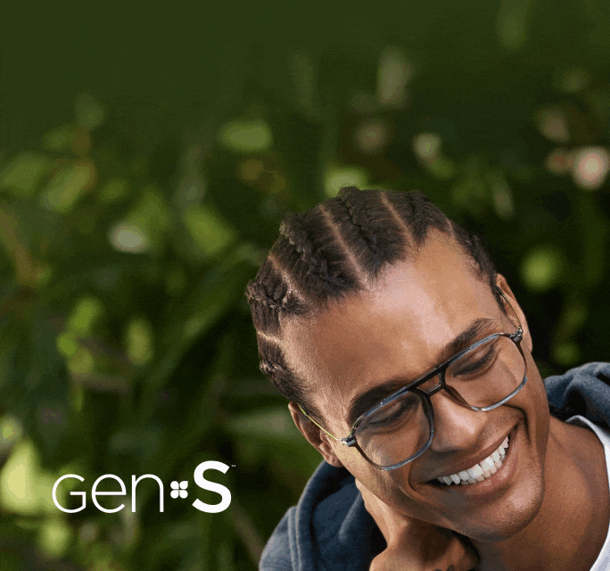 Homme souriant aux cheveux tressés portant des lunettes navigateur en gris translucide avec des verres foncés teintés Gen S, à côté du logo « Gen S » sur fond de verdure extérieure floue.