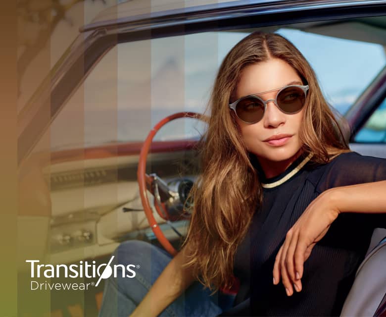 A woman sitting in the front seat of a car wearing round glasses with Transitions lenses that change from dark to darker.