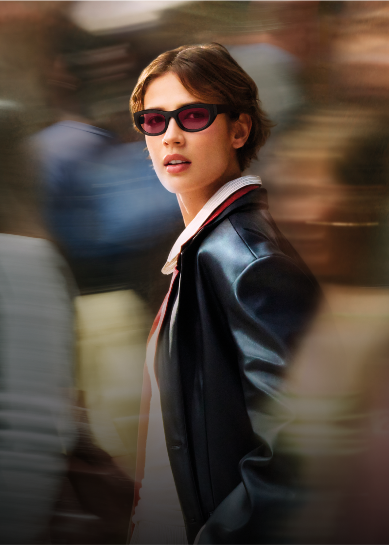 Woman with short brown hair wearing narrow black sunglasses with pink-tinted lenses. They are dressed in a black leather jacket over a collared shirt and are looking slightly to the side. The blurred background suggests a busy urban scene, giving the photo a sense of motion and energy.