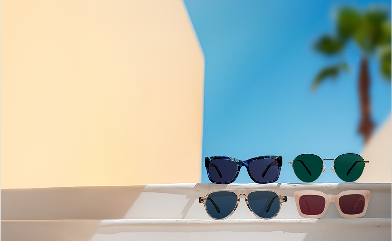 Four pairs of sunglasses are neatly arranged on white steps under bright sunlight, with a pastel peach wall and a clear blue sky in the background, suggesting a warm and stylish summer setting.. The frames vary in color and shape.