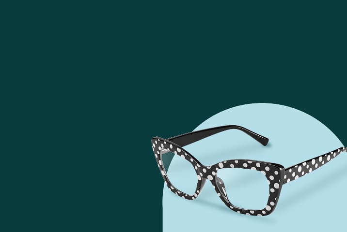 Black cat-eye eyeglasses with white polka dots resting on a light blue oval platform against a dark green background.