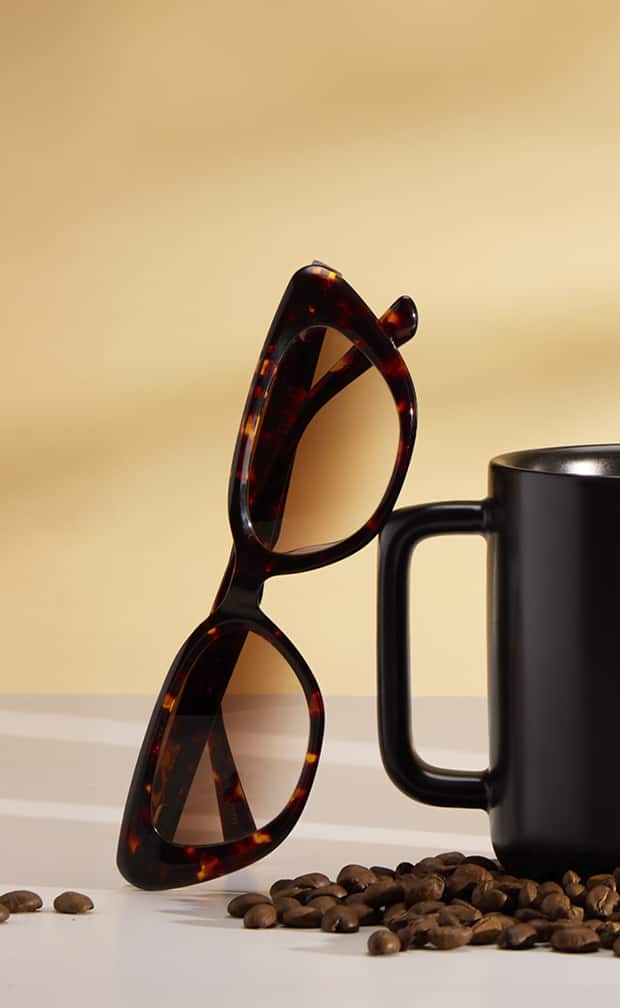 Zenni tortoiseshell sunglasses next to a black coffee mug and coffee beans on a tan background.