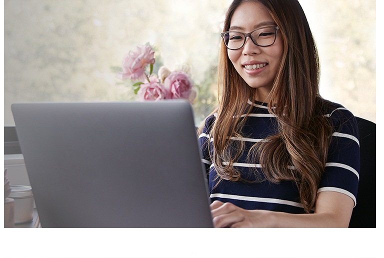 A young woman with long brown hair wearing Blokz by Zenni blue blocker glasses smiles while using her laptop computer.