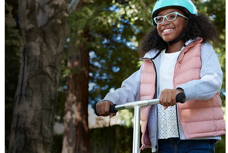 Young girl smiles while riding a scooter outside wearing medium-sized Spark cat-eye glasses # 4440316 in sky blue from the Style Squad kids’ collection with Blokz Trivex lenses and matching light blue helmet.