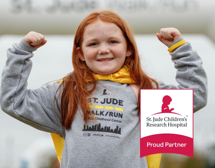 A girl with long red hair wearing a gray St. Jude sweatshirt and a yellow cape, holding up her arms in a gesture of strength. The red and white logo of St. Jude's Children's Research Hospital appears in the bottom right corner, with the text: Proud Partner.