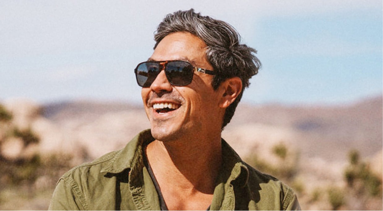 Man wearing khaki green jacket and sunglasses smiles towards the sun with Joshua Tree National Park in background.