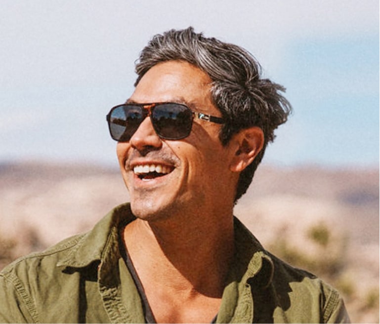 Man wearing khaki green jacket and sunglasses smiles towards the sun with Joshua Tree National Park in background.