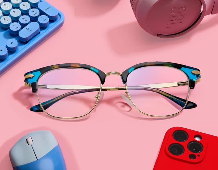Lentes de tortuga con cejas Zenni sobre un escritorio rosa con fonos, teclado, mouse y teléfono.