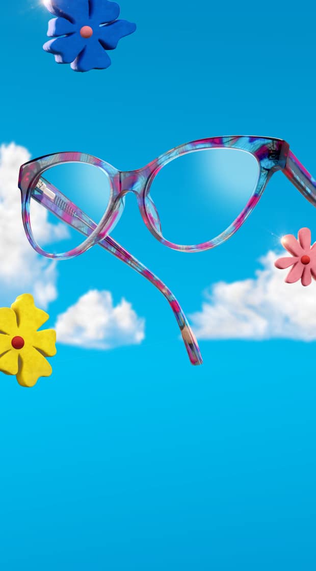 Multicolored floral-patterned cat-eye eyeglasses floating against a bright blue sky with soft white clouds and colorful clay flowers.
