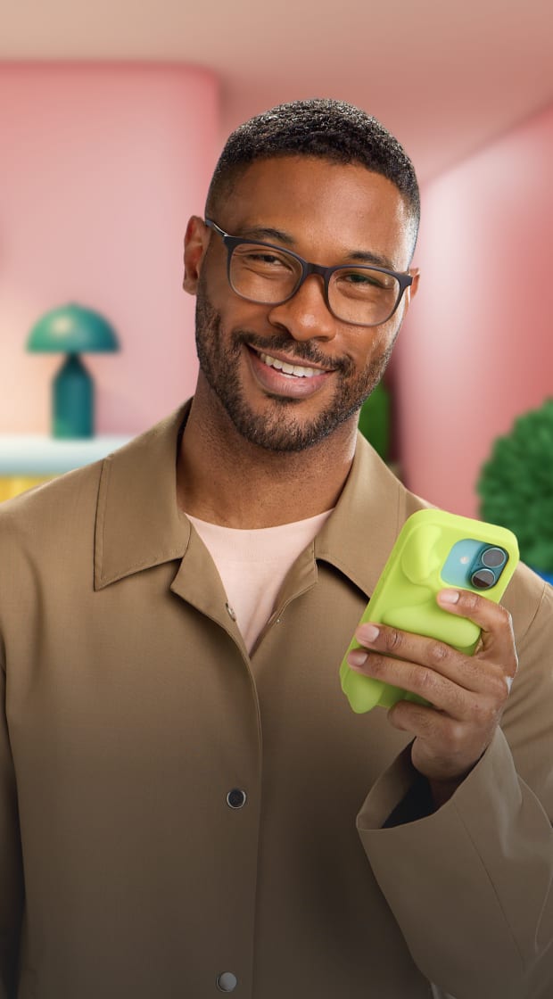 Hombre sonriente con barba corta usando lentes rectangulares oscuros y una camisa de botones de color marrón claro, sosteniendo un teléfono inteligente verde limón en una habitación brillante y colorida con paredes rosas y muebles minimalistas azules y amarillos.