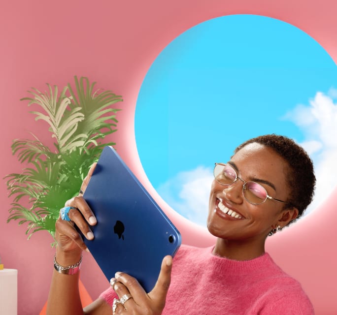 A smiling young woman with short hair holding a dark blue tablet, wearing thin metal wireframe eyeglasses with a pinkish reflection and a fuzzy pink sweater, set against a pink wall with a circular window opening to a blue sky.