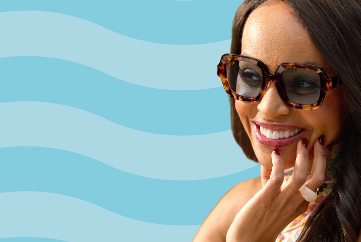 Close-up of a smiling woman with dark hair touching her chin while wearing oversized square tortoiseshell sunglasses, set against a light blue background with abstract darker blue waves.