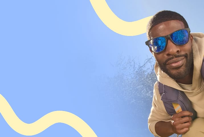 A man with close cropped black hair wearing black square Zenni sports sunglasses with blue mirror lenses. Blue sky and trees background with a yellow squiggle motif.