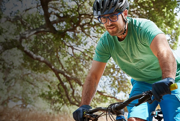 Tony on his mountain bike outdoors wearing translucent blue Olvera square glasses #449416 with Blokz Trivex lenses, a bike helmet, green t-shirt, and blue shorts.