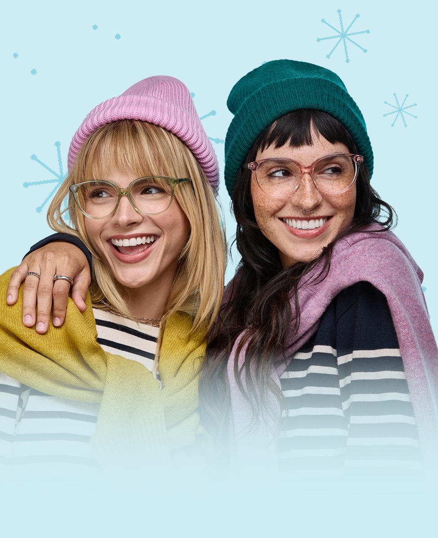 A blond woman wearing clear green glasses and a pink knit hat being hugged by a brunette woman wearing clear pink glasses and a green knit, on a blue background with a star or snowflake motif.