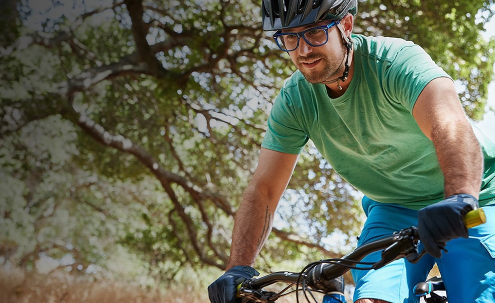 Tony on his mountain bike outdoors wearing translucent blue Olvera square glasses #449416 with Blokz Trivex lenses, a bike helmet, green t-shirt, and blue shorts.