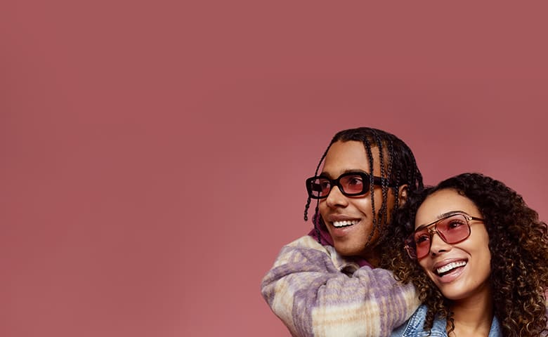 A happy couple, man with braids and woman with curly hair, both wearing Zenni FL-41 rose-tinted glasses and denim attire.
