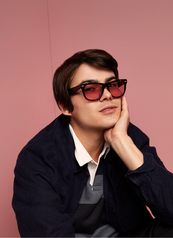 A thoughtful man in a dark jacket and white collared shirt wearing oversized Zenni FL-41 rose-tinted glasses, seated against a pink wall.
