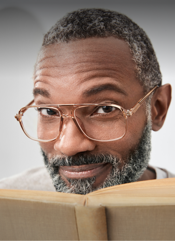A black man with salt and pepper hair wearing aviator reading glasses and holding a book smiles with his eyebrows raised.