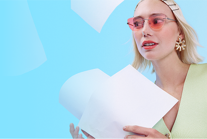 Blonde woman in peach-tinted rimless glasses with white paper sheets flying around her on a blue background.