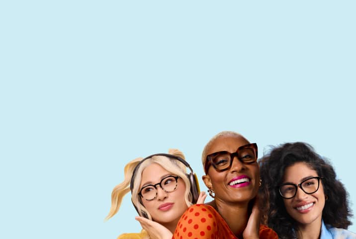 Group portrait of three smiling women wearing stylish eyeglasses against a light blue background, including a blonde woman with headphones in round spotted frames, a woman with short blonde hair in thick square tortoiseshell frames, and a woman with curly dark hair in black rectangular frames.